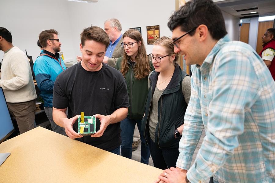 A young man in a black shirt holds a green cube with yellow attennae in his hands while a woman in glasses and a man in a plaid shirt look on.