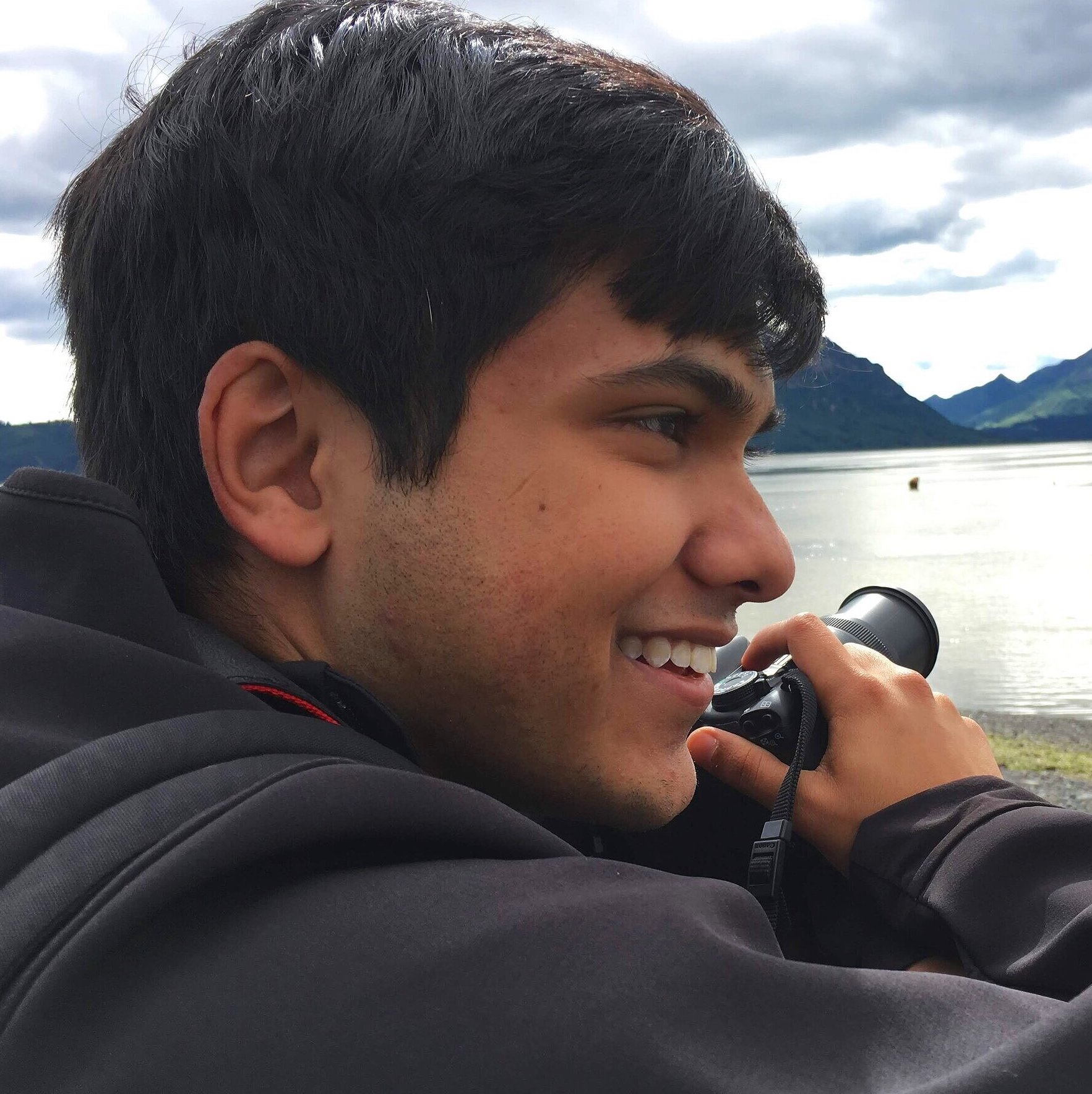 Photo of Mihir Bala holding a camera in front of water and mountains