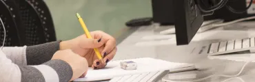 person holding a writing implement using a notepad, sitting in front of a computer